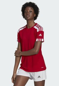 Rotes Sportshirt mit weißen Streifen an den Schultern und Adidas-Logo auf dem Ärmel; kombiniert mit weißen Shorts. Glattes Material, reguläre Passform.