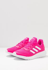 Un par de zapatillas de跑 de Adidas en un vibrante color rosa fucsia con franjas blancas y suelas blancas, sobre una superficie blanca contra un fondo gris claro.