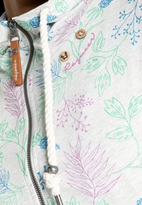 Ljust grå hoodie med ett blommönster i blått, grönt och lila. Har en dragkedja med en läderdragknopp och knutna repdragningar.