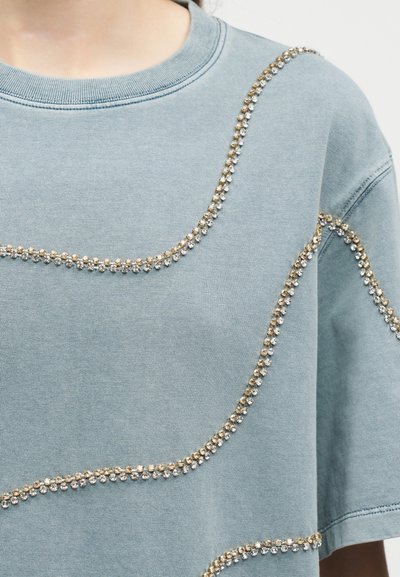 Ανοιχτό μπλε πουκάμισο με κοντά μανίκια, διακοσμημένο με κυρτές γραμμές από χρυσές και ασημί στρας στο μπροστινό μέρος και στα μανίκια.