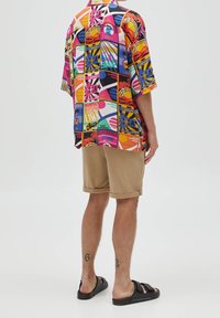 Kurzarmhemd mit hellen, bunten Grafikmustern, kombiniert mit beigen Shorts. Schwarze Slipper runden den lässigen Look ab.