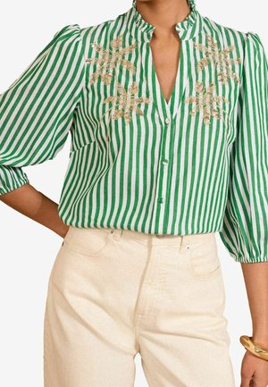 Zielona i biała bluzka w pionowe paski z złotym kwiatowym haftem, rękawy 3/4 z bufkami, w zestawieniu z beżowymi, wysokim stanem spodniami i złotą bransoletką.