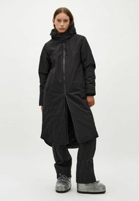 Zwarte lange jas met een capuchon, voorzien van een rits aan de voorkant en zijzakken. Draag over gewatteerde zwarte broek en glanzende zilveren platform schoenen.