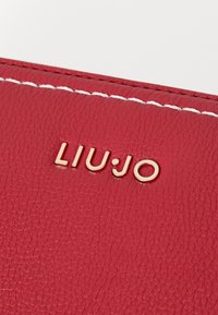 Portafoglio in pelle rossa texture con cuciture bianche e logo dorato "LIUJO". La vista ravvicinata mette in evidenza i dettagli del materiale e del marchio.