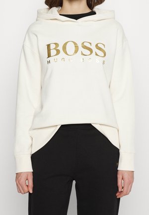 Nainen valkoisessa hupullisessa paidassa, jossa on kultaiset "BOSS HUGO BOSS" -tekstit, ja mustissa housuissa, seisomassa yksinkertaisen vaalean taustan edessä.