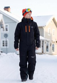 Schwarzer Schneeanzug mit orangen Akzenten, Frontreißverschluss, Reißverschlusstaschen, Kapuze und Handschuhen. Getragen mit einer leuchtend orangefarbenen Mütze und Skibrille.