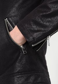 Svart läderjacka med en blank yta, prydd med silverfärgade dragkedjor och fickor på sidorna, som visar en närbild av tygstrukturen.