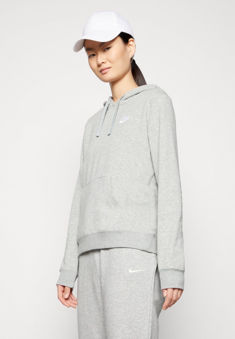 Grauer Hoodie mit einer vorderen Tasche und einem weißen Nike-Logo auf der Brust. Kombiniert mit grauen Jogginghosen und einer weißen Kappe.