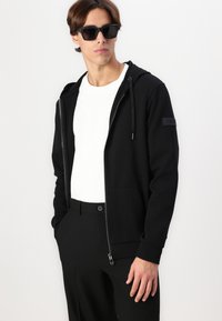 Crna zip-up dukserica s prednjim džepom, kapuljačom sa šniranjem i teksturiranim logom na rukavu, nosi se preko bijele majice i crnih hlača.