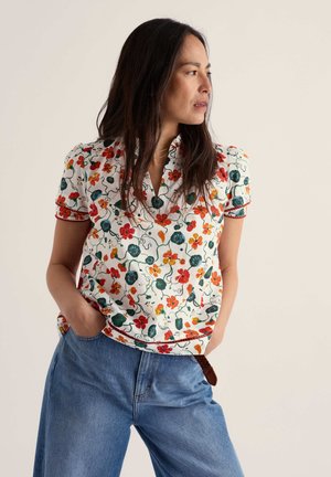 Mujer de pie con las manos en los bolsillos de los vaqueros, lleva una camiseta blanca de manga corta con estampado floral en rojo, naranja y verde, mirando hacia la derecha.