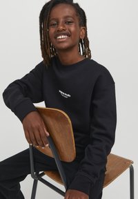 Felpa nera con scollatura rotonda e logo in bianco sul petto, abbinata a pantaloni scuri. Tessuto testurizzato e vestibilità rilassata.