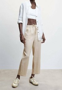 Beige linnen broek met een trekkoord in de taille, gecombineerd met een korte witte tanktop en een losse witte overhemd. Lichtgekleurde sneakers.