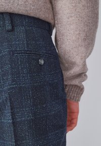 Pantalones texturizados azul marino con un sutil patrón de cuadros, botón de tela y un bolsillo trasero. Suéter tejido beige visible en la parte superior.