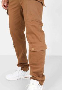 Bruine cargobroek met meerdere utiliteitszakken, gemaakt van duurzaam materiaal; voorzien van knopen op de zakken en geribde enkelmanchetten. Draagt samen met witte sneakers.