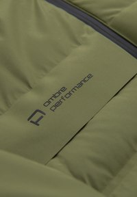 Gros plan sur un tissu matelassé vert olive avec une fermeture éclair noire et un logo "ombre performance" cousu sur la surface.