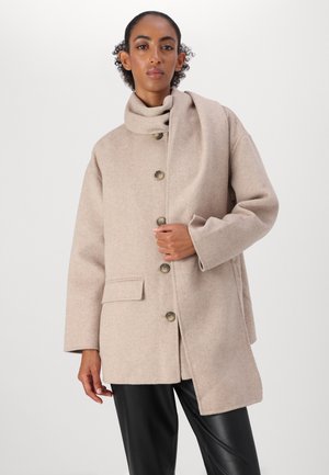 Cappotto in misto lana beige con colletto alto, bottoni frontali, tasche laterali e vestibilità relaxed, abbinato a pantaloni neri lucidi.