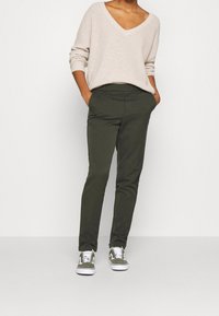 Beige stickad tröja med V-ringning; olivgröna slim-fit byxor; grå och vita sneakers med gröna accenter; händerna i fickorna.