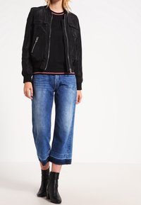 Donna che indossa una giacca bomber nera con zip, una maglietta nera con rifiniture rosse, jeans blu a gamba larga e stivaletti neri.