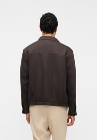 Άνδρας με κοντά σγουρά μαλλιά, που φοράει σκούρο καφέ μπουφάν και ανοιχτό μπεζ παντελόνι, στέκεται με την πλάτη προς την κάμερα σε λευκό φόντο.