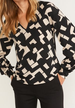 Kobieta w czarno-beżowej bluzce z abstrakcyjnym wzorem, z długim rękawem i dekoltem w serek, z rękami w kieszeniach, w połączeniu z czarnymi spodniami.