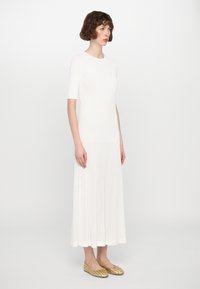 Robe blanche longueur genou avec des manches courtes, texture côtelée au niveau de la jupe, haut lisse, complétée par des ballerines tressées dorées.