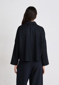 Schwarze Nadelstreifenjacke mit einem cropped Design, weiten Ärmeln und einem Kragen. Der Stoff sieht strukturiert aus und wird mit dunkel, weiten Hosen kombiniert.