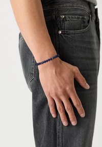 Perlenarmband aus dunkelblauen Steinen mit glänzenden silbernen Akzenten, getragen am Handgelenk neben dunkelgrauen Jeans.