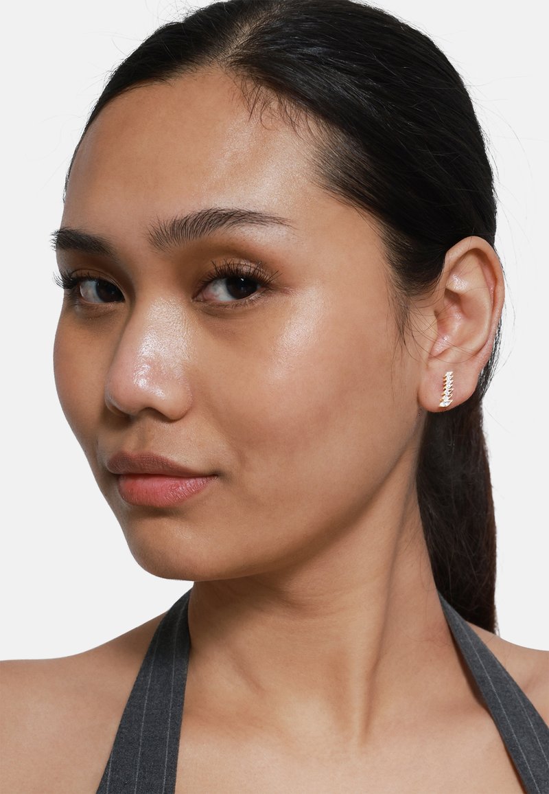 Orecchini dorati con design a zigzag. Il modello ha una pelle liscia, capelli scuri raccolti e indossa un top grigio a spalle scoperte.
