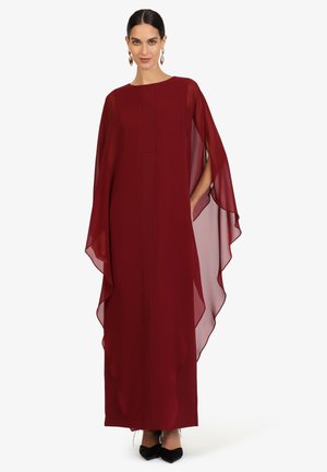 Frau trägt ein langes, locker sitzendes Kleid mit transparenten Ärmeln in Bordeaux, schwarze Schuhe und goldene Creolen, steht auf weißem Hintergrund.