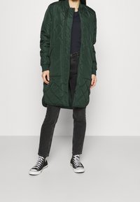 Tmavě zelený prošívaný dlouhý kabát s límcem a druky, stylizovaný přes černý top, spárovaný s černými džíny a teniskami s vysokou podešví.