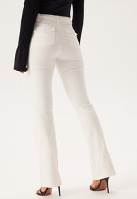 Pantalons blancs évasés avec deux poches arrière et une finition lisse, associés à un haut à manches longues noir, mettant en valeur une silhouette élancée.