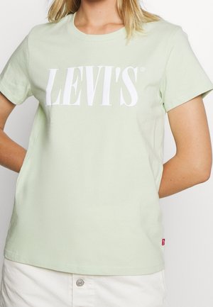 Kobieta ubrana w jasnozieloną koszulkę z krótkim rękawem z dużym białym logo "LEVI'S" na piersi oraz białą spódnicę na guziki.