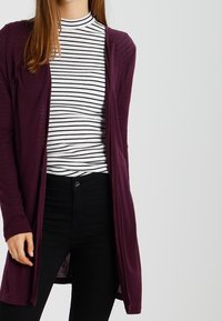 Lång, öppen framsides cardigan i djupt lila tyg. Bärs över en svart-vit randig polotröja och svarta jeans. Mjuk textur med ärmar.