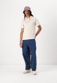 Beige cotton t-shirt with a small logo on the chest, paired with loose-fitting blue denim jeans and light-colored sneakers. Accented by a lavender bucket hat.