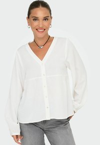 Biely dlhý rukáv blouse s V-výstrihom, predným zapínaním na gombíky a detailom s nariaseným pásom. Mäkka tkanina s hladkou textúrou. Nosené cez šedé džínsy.
