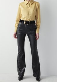 Sorte faux læder flared bukser med sømdetaljer, parret med en gylden skjorte med knapper og et bredt sort bælte med en guldbuckle.