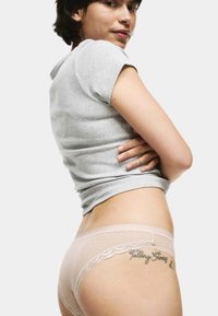 Vrouw in een grijze T-shirt met korte mouwen en doorschijnende roze onderbroek met kant, die een "Verhalen Vertellen" tatoeage op haar heup toont.