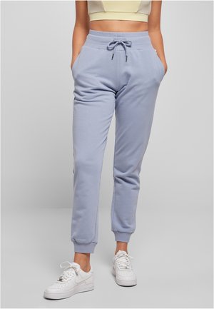 Femme portant un pantalon de jogging bleu clair à cordon et des baskets blanches, debout les mains dans les poches sur un fond uni.