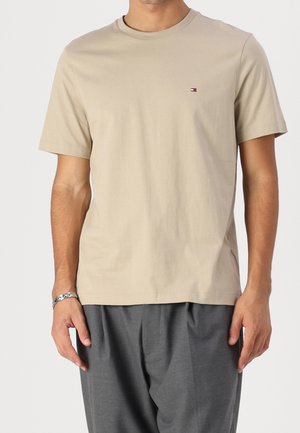 Mężczyzna ubrany w beżowy t-shirt z krótkim rękawem z małym logo na piersi, w połączeniu z ciemnoszarymi spodniami i srebrną bransoletką na lewej ręce.
