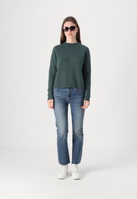 Pull en tricot vert avec un col rond, associé à un jean bleu clair et des baskets blanches. Le mannequin porte des lunettes de soleil noires et se tient droit.