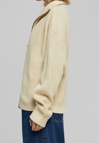 Béžový pletený kardigan s texturovaným vzorem, žebrovanými lemy a límcem, vybavený zipem a volným střihem. Nošený přes tmavě modrou džínovou sukní.