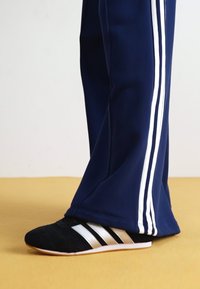 Navy athletic pants with three white side stripes, paired with black suede shoes featuring three silver diagonal stripes and white sole.