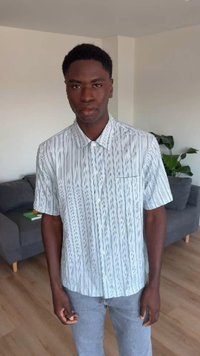 Νέος άνδρας με κοντά σγουρά μαλλιά που φοράει ένα λευκό σύντομο πουκάμισο με σχέδια και ανοιχτό γκρι παντελόνι, στέκεται σε εσωτερικό χώρο.