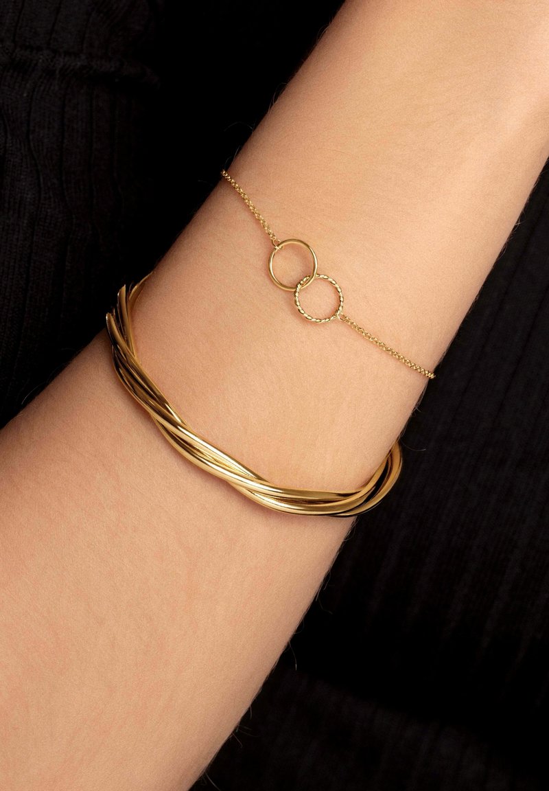 Deux bracelets en or au poignet, une fine chaîne avec deux anneaux entrelacés, l'autre un design torsadé plus épais.