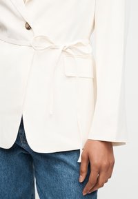 Γυναίκα που φοράει λευκό σακάκι με λεπτή ζώνη γραβάτα και πτερύγιο τσέπης, σε συνδυασμό με μπλε τζιν, με το χέρι χαλαρό στο πλάι.