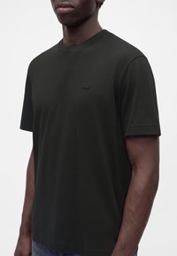 T-shirt noir à manches courtes avec col rond et logo brodé discret sur la poitrine, porté par une personne à la peau foncée et aux cheveux courts.