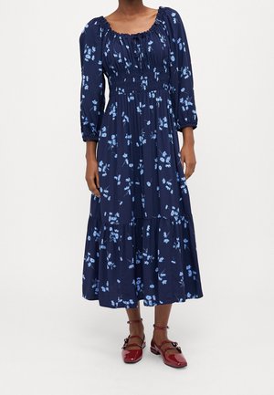 Marineblauwe midi-jurk met lichtblauwe bloemenprint, poflange mouwen, gerimpelde taille en geplooide zoom, gecombineerd met rode lak Mary Jane-schoenen.