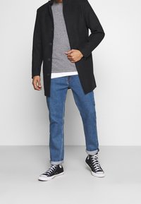 Černý kabát na zapínání přes knoflíky přes šedý svetr, modré džíny a černé tenisky s bílými podešvemi. Hladká tkanina, moderní design, neformální střih.