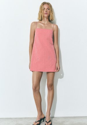 Ung kvinde står op ad en hvid væg iført en rød- og hvidternet minikjole med spaghettistropper og sorte sandaler med hæl.