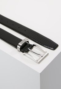 Schwarzer Ledergürtel mit glatter Oberfläche, einer silbernen rechteckigen Schnalle mit Dorn und mehreren Löchern zur Größenanpassung.